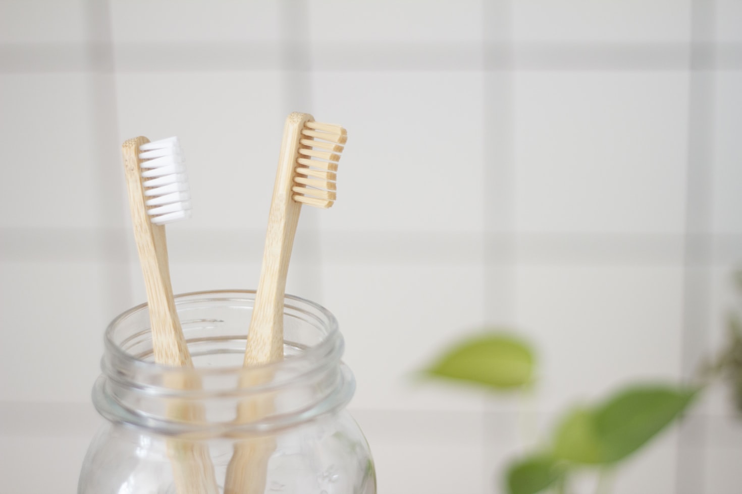 Two bamboo toothbrushes in a glass jar, one with white bristles and the other with beige bristles, against a tiled wall background.